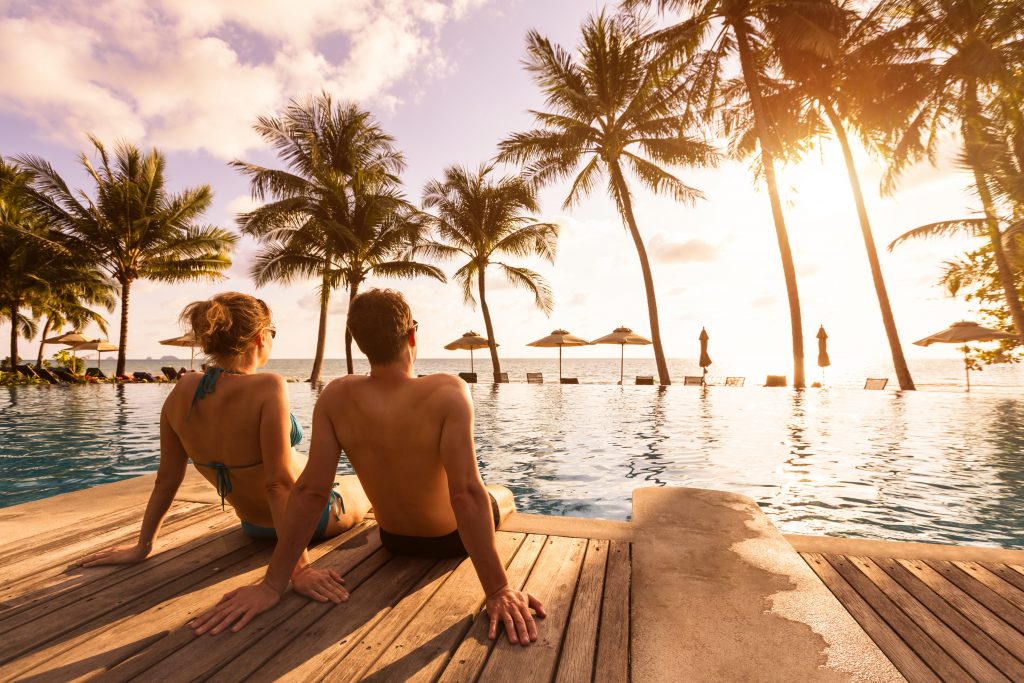 Couple sitting by a tropical resort pool at sunset overlooking palm trees and the ocean.